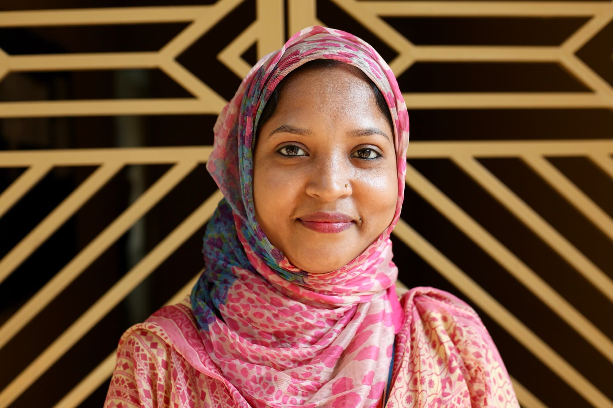 A woman in a bright headscarf smiling in front of a gold geometric screen